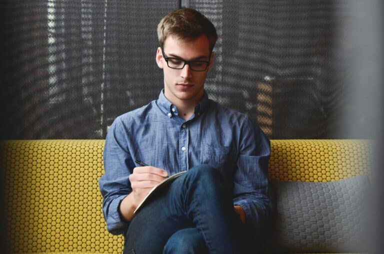 Un jeune homme portant des lunettes écrit dans un cahier assis.